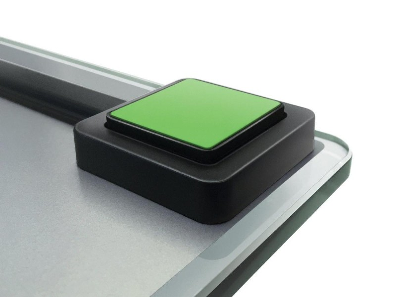 Primer plano de un botón cuadrado verde en una báscula, marco negro, superficie plateada.