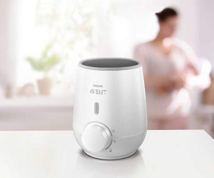 Un calienta biberones Philips Avent blanco con dial y botón. Una mujer borrosa en el fondo.