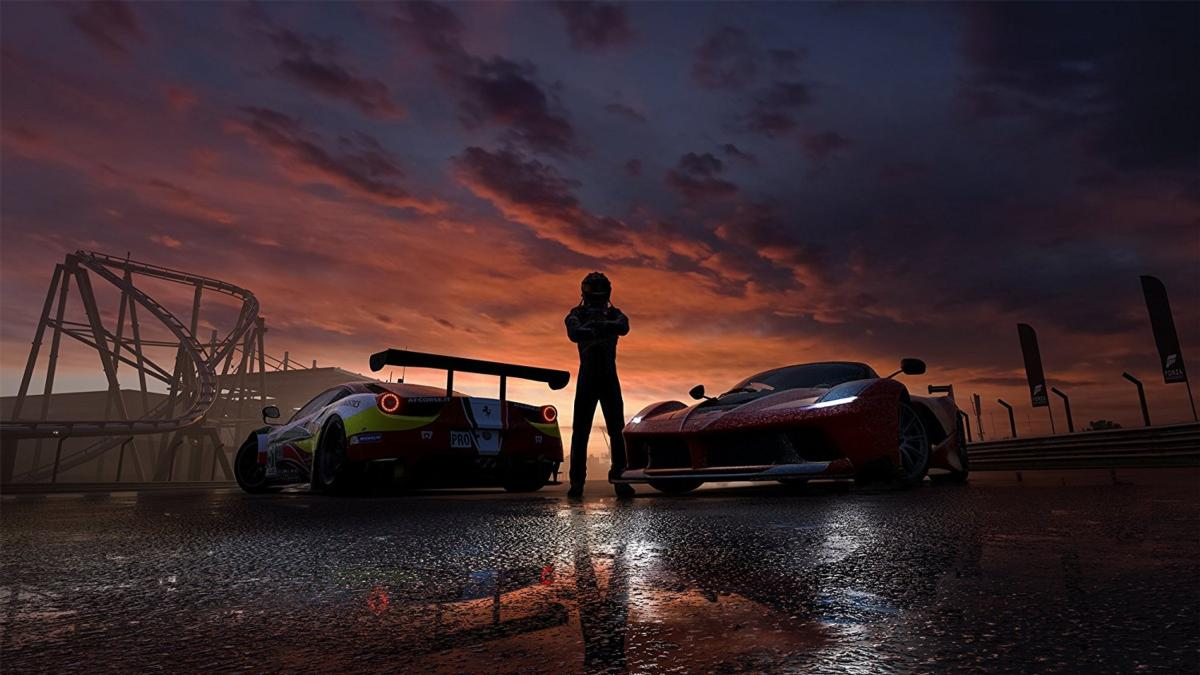 Una persona está de pie entre dos coches de carreras en una pista mojada al atardecer. Una montaña rusa en el fondo.