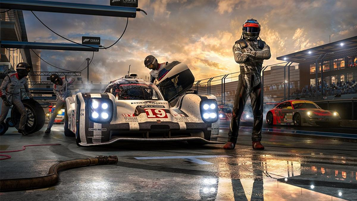 Coche de carreras en boxes, pilotos y equipo preparándose. Un piloto está con los brazos cruzados. Otro coche corre en el fondo.