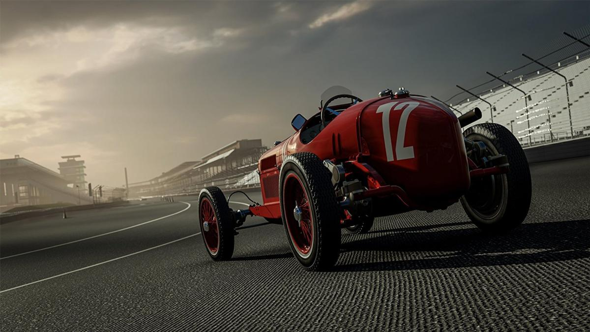 Un coche de carreras rojo vintage con el número 12 en el lateral, en una pista. El fondo muestra una tribuna y un cielo nublado.