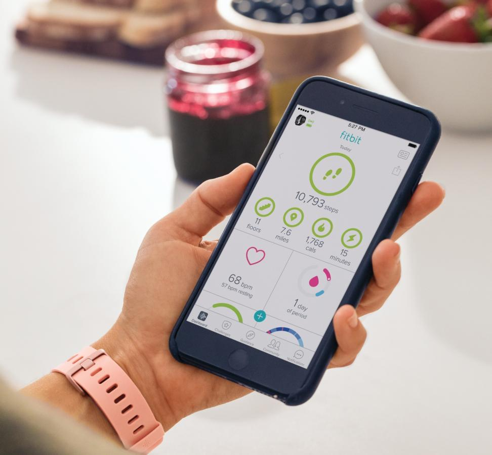 Mano sosteniendo teléfono mostrando aplicación de fitness con pasos, millas y calorías. Pulsera rosa.