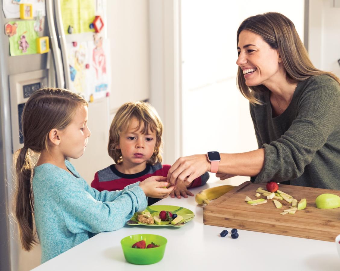 Una mujer y dos niños están en una mesa. La mujer corta fruta, los niños comen. Ambiente de cocina.