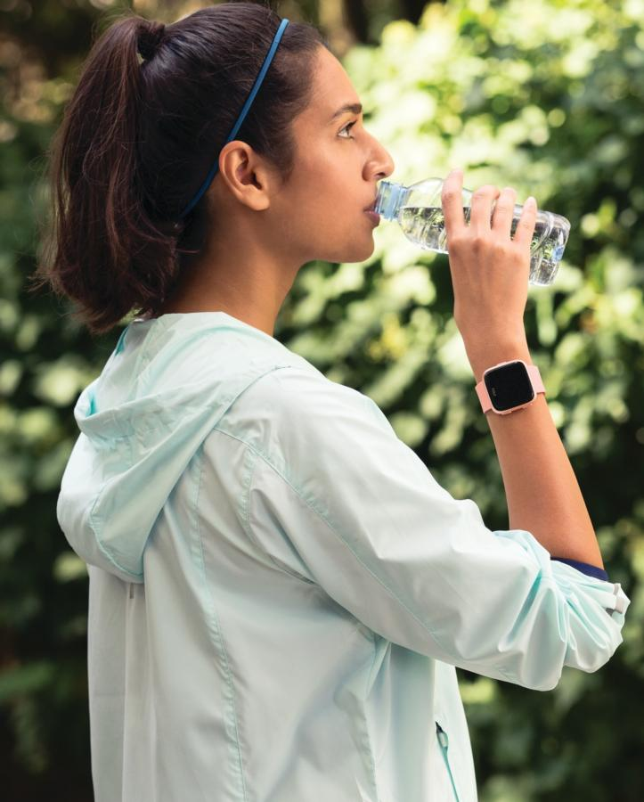 Mujer bebiendo agua de una botella, smartwatch, parque, vista lateral.