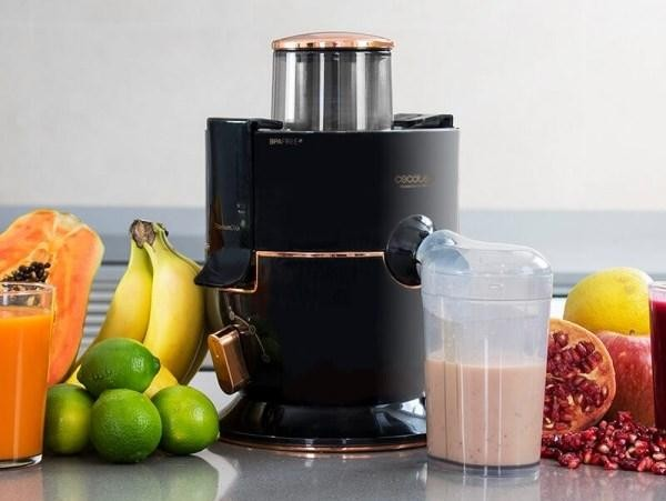 Una licuadora negra y cobre con frutas, vaso de jugo y vaso de batido.