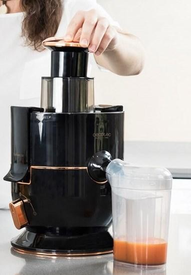 Persona usando una exprimidora negra y cobriza. Un recipiente transparente recoge el jugo.