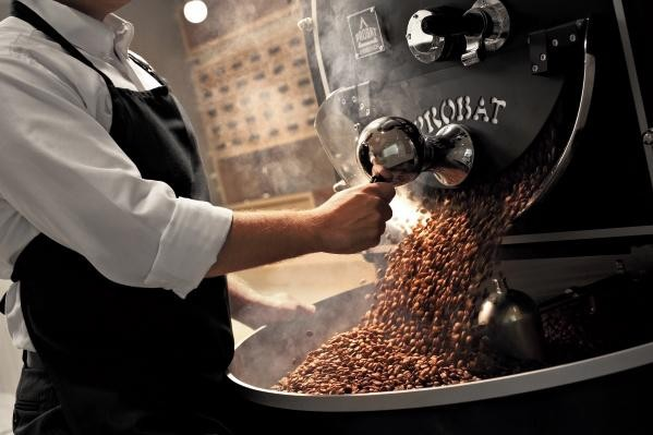 Una persona con camisa blanca y delantal negro vierte granos de café tostados en una máquina.