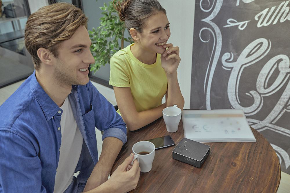 Ein Mann und eine Frau an einem Tisch mit einer Kaffeetasse und einem kleinen schwarzen Projektor.