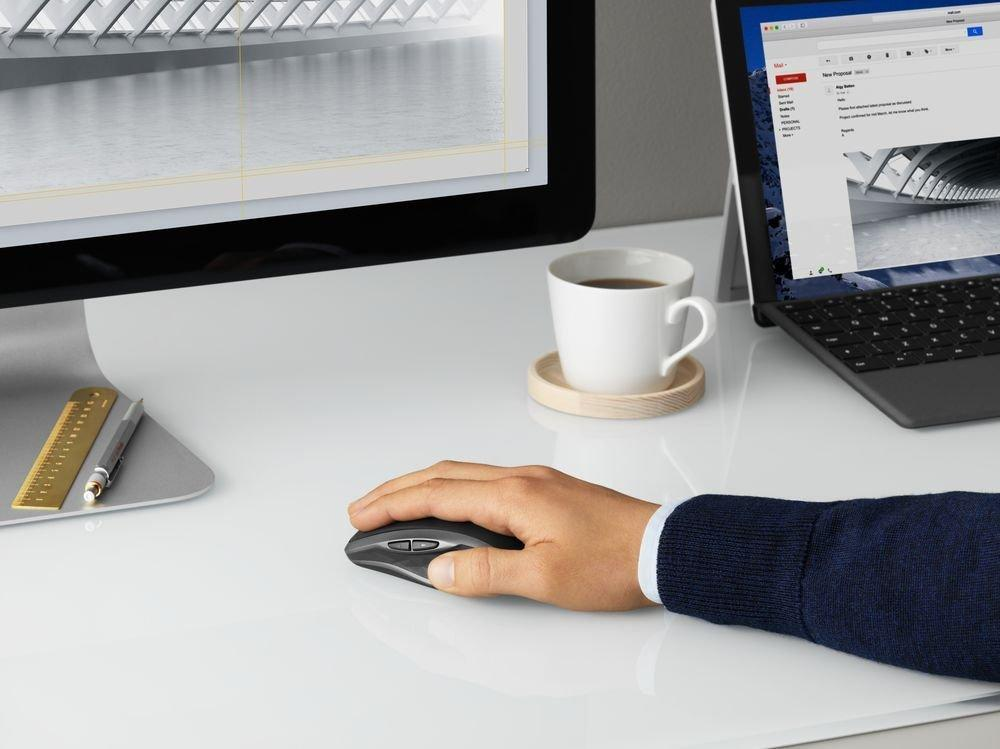 Una mano usando un ratón en un escritorio blanco. Monitor, portátil, taza y posavasos visibles.