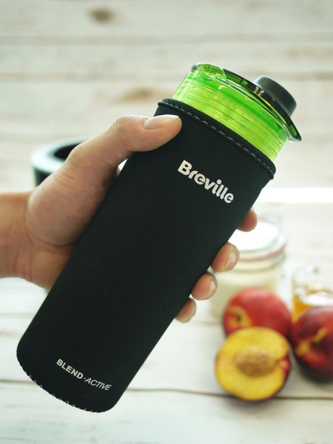 Mano sosteniendo una botella negra Breville Blend Active con contenido verde. Duraznos visibles al fondo.