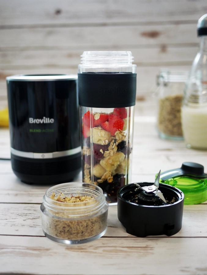 Una licuadora con ingredientes en un vaso. Tarros y partes de la licuadora también en la superficie de madera.
