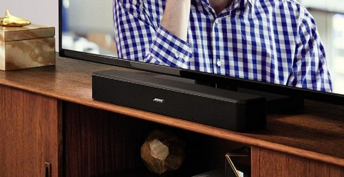 Una barra de sonido negra debajo de un televisor, sobre un mueble de madera. TV muestra persona.