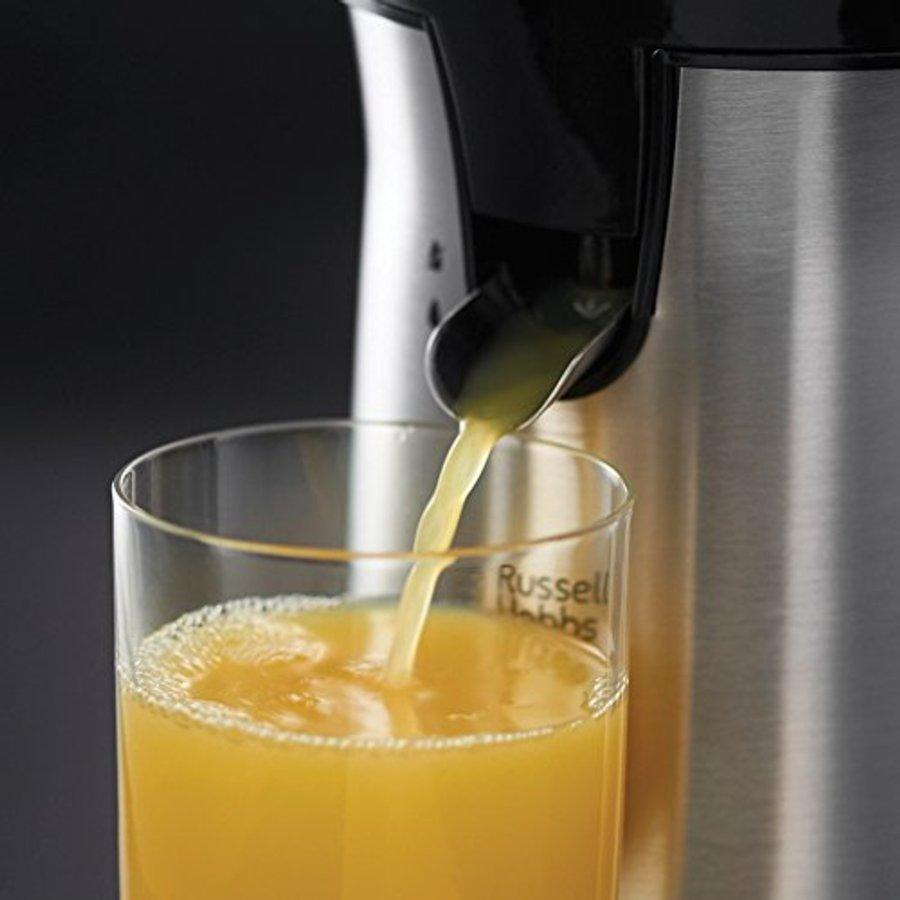 Jugo de naranja vertiéndose de una exprimidora a un vaso. La exprimidora es metálica.