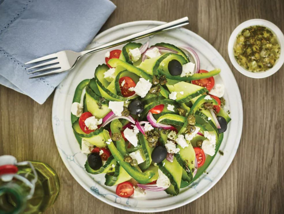 Salade dans une assiette avec concombres, tomates, olives et fromage. Fourchette et vinaigrette à côté.