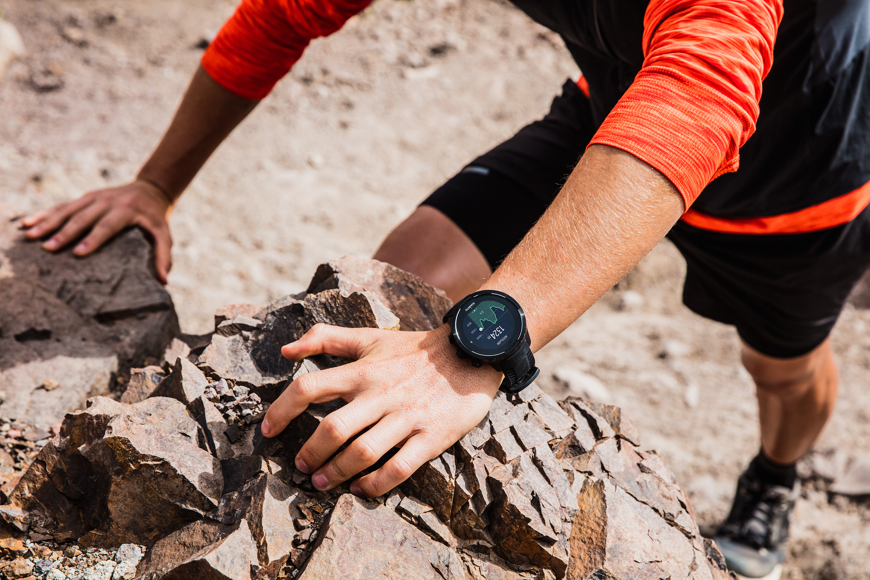 Mann klettert auf Felsen, trägt schwarze Shorts, orangefarbenes Hemd und schwarze Uhr.