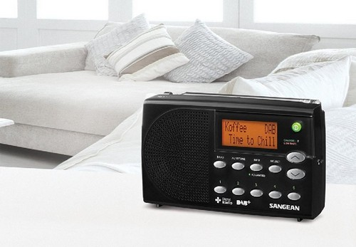 Radio digital negro con pantalla naranja, colocado cerca de almohadas en un sofá blanco.