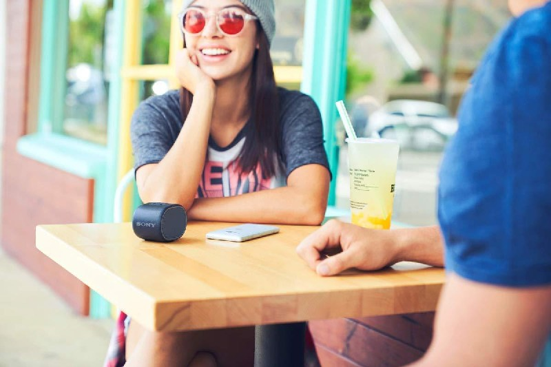 Una mujer con gafas de sol y un hombre están sentados en una mesa. Un altavoz está en la mesa.