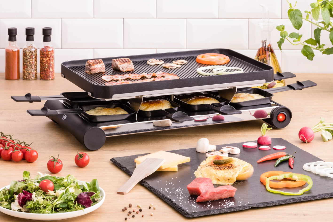 Raclette grill with food, including meat, vegetables, cheese, and bread. The table is wooden, and there is a white wall.