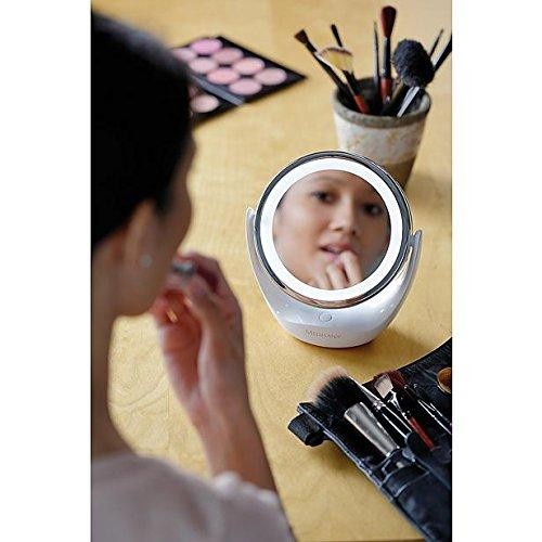 Una mujer mirándose en un espejo de maquillaje iluminado. Se ven brochas y suministros de maquillaje.