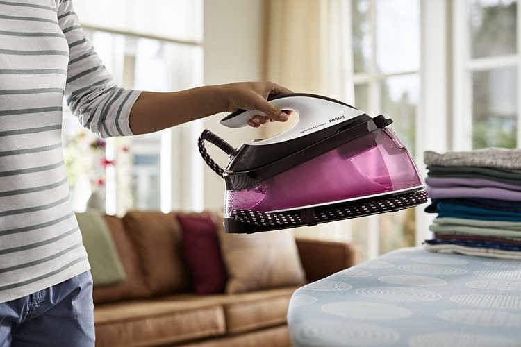 Mujer sosteniendo plancha cerca de ropa. Tabla de planchar y sofá visibles.