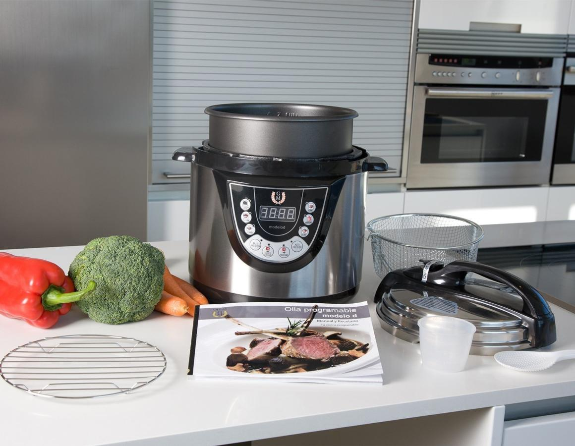 Una olla a presión plateada y negra con verduras y un libro de recetas sobre una superficie blanca.