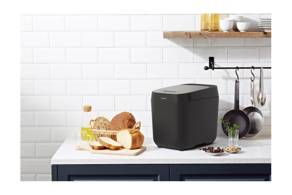 Ein schwarzer Brotbackautomat auf einer weißen Arbeitsplatte mit Brot, Öl und anderen Küchenutensilien im Hintergrund.