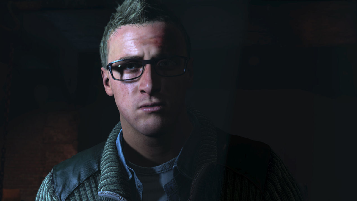 Ein Mann mit Brille in einem schwach beleuchteten Raum, der den Betrachter ansieht. Er trägt einen Pullover und eine Jacke.