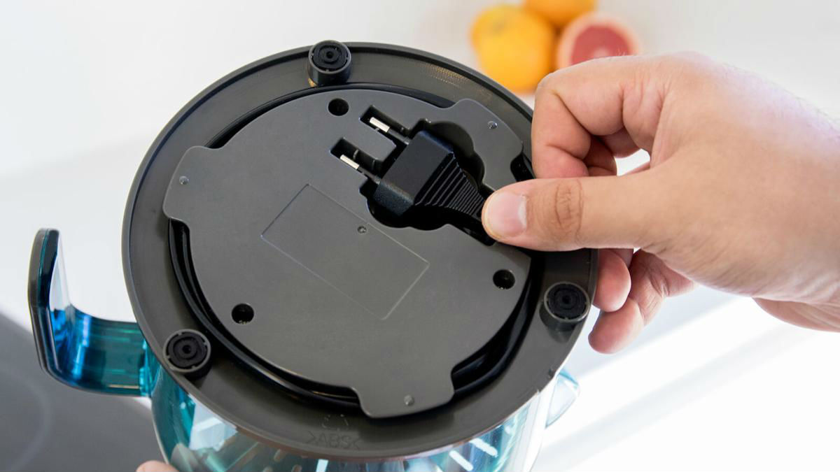 Primer plano de una mano enchufando un cable de alimentación negro en la base de un electrodoméstico de cocina azul y transparente.