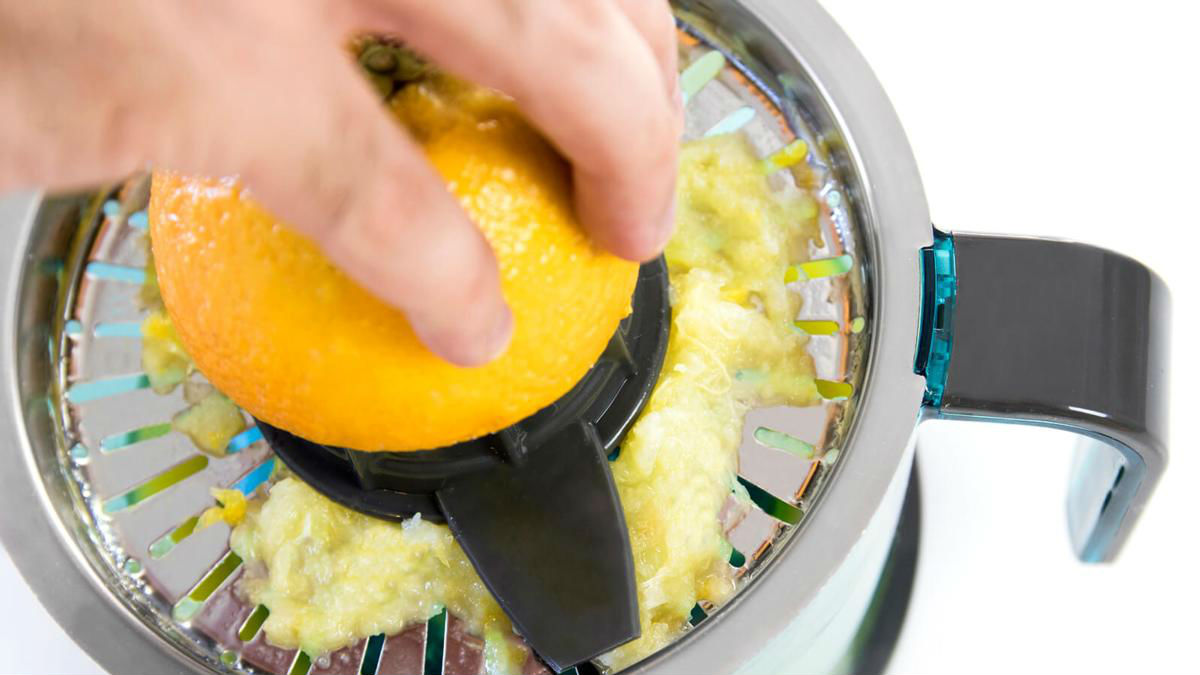 Una mano exprimiendo una naranja en una exprimidora, extrayendo jugo. La exprimidora es de metal y tiene un colador.