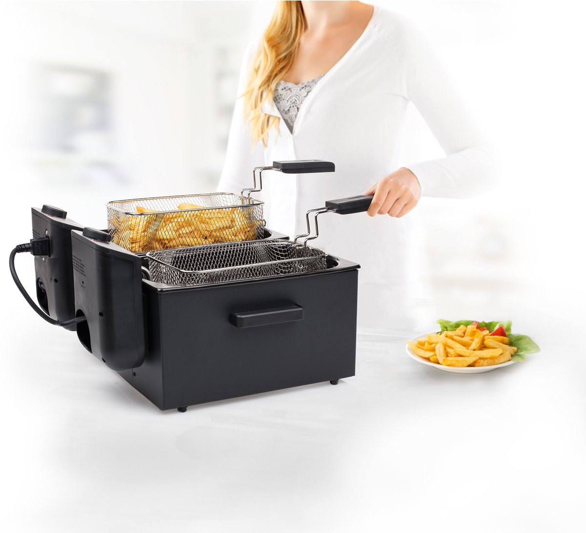 A woman frying french fries in a black fryer, with a plate of fries on the side.