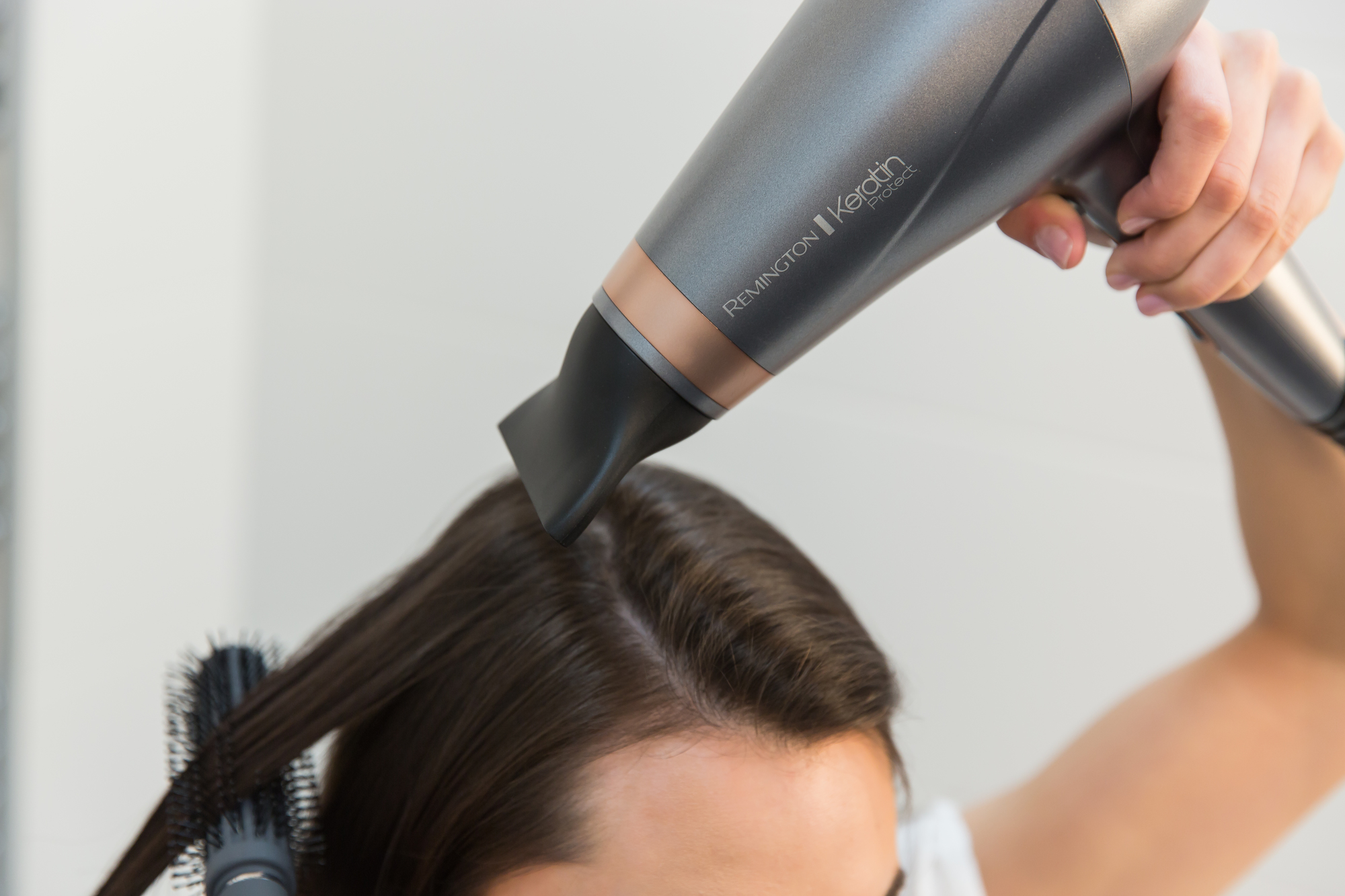 A person is drying hair with a gray Remington Keratin hairdryer and a round brush.