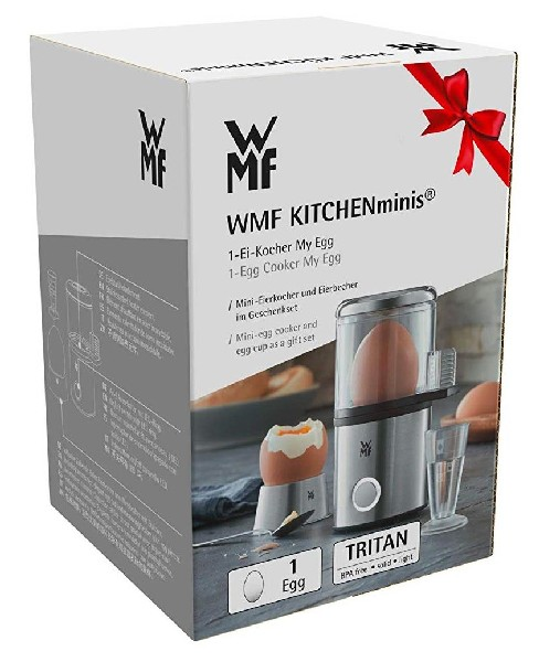 Caja con la cocinadora de huevos WMF Kitchenminis, un lazo rojo y un huevo en un soporte. El fondo está borroso.