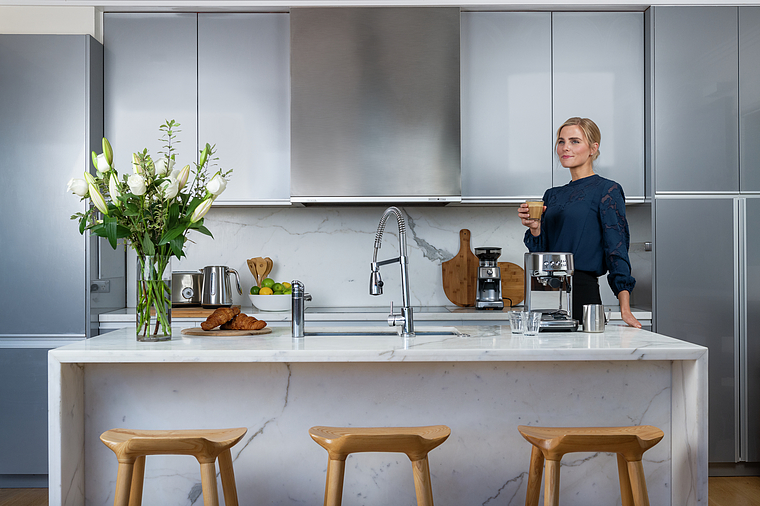 Frau in Küche mit Blumen, Espressomaschine und Croissants. Moderne Küche mit Marmorinsel.