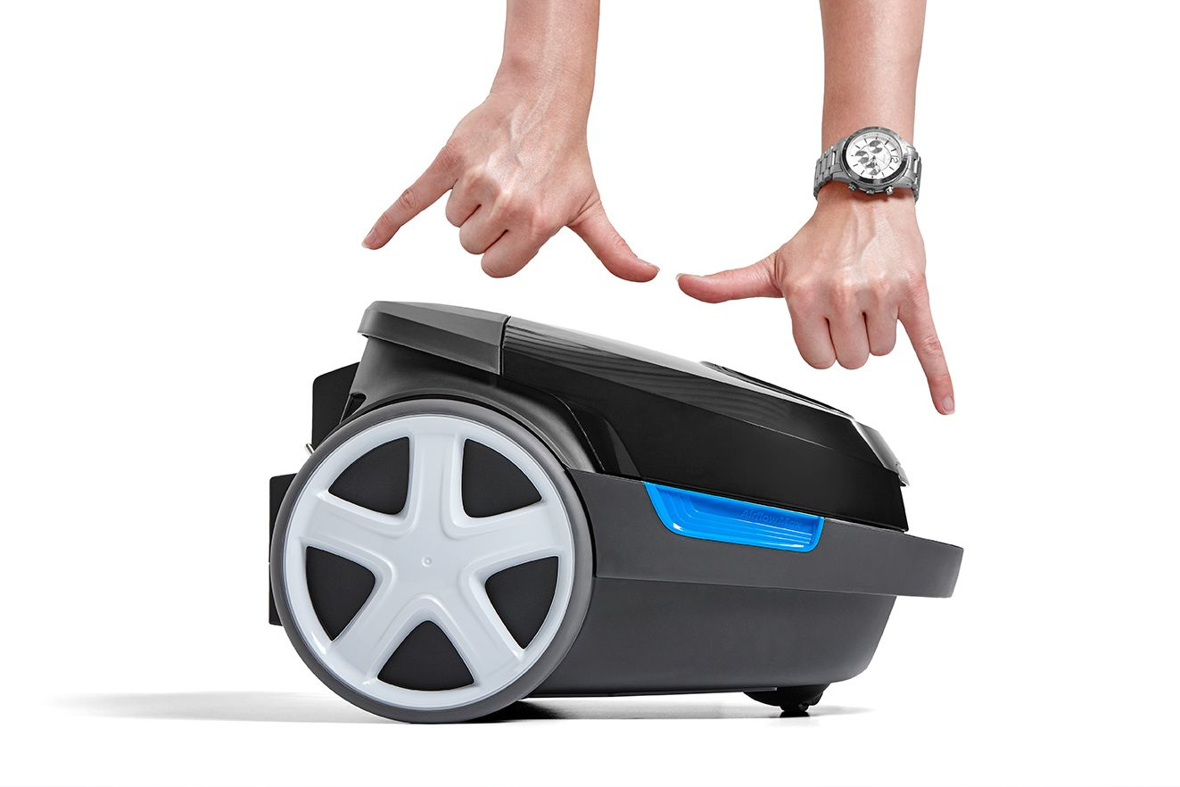 Hände zeigen auf einen schwarzen Staubsauger mit weißen Rädern. Ein Handgelenk hat eine Uhr. Blaue Akzente an der Seite.