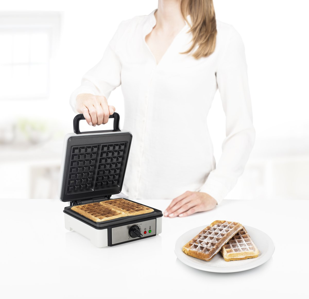 Persoon die een wafelijzer vasthoudt met twee wafels en een bord met wafels op een wit oppervlak.