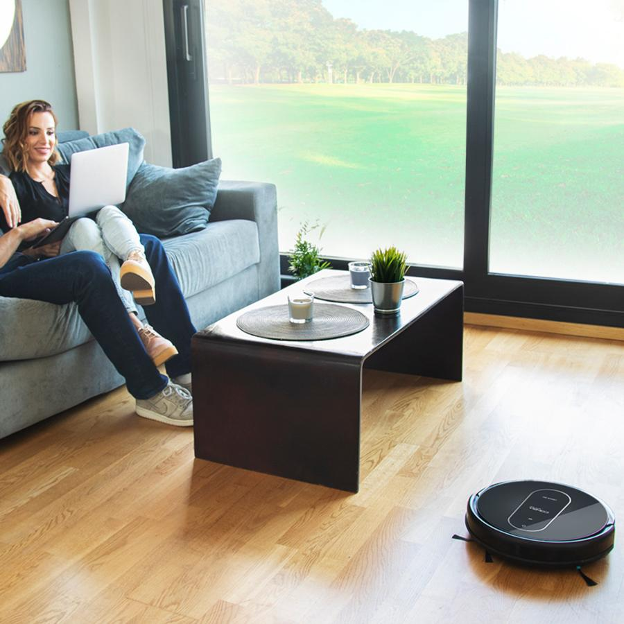 Una pareja en un sofá con un portátil. Una aspiradora robot está en el suelo. El fondo es una ventana.