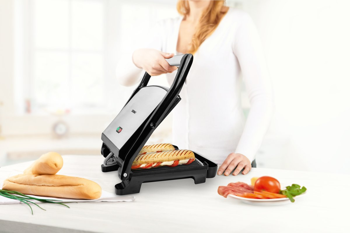 Sandwich maker with sandwiches inside, woman holding handle, on table with food.
