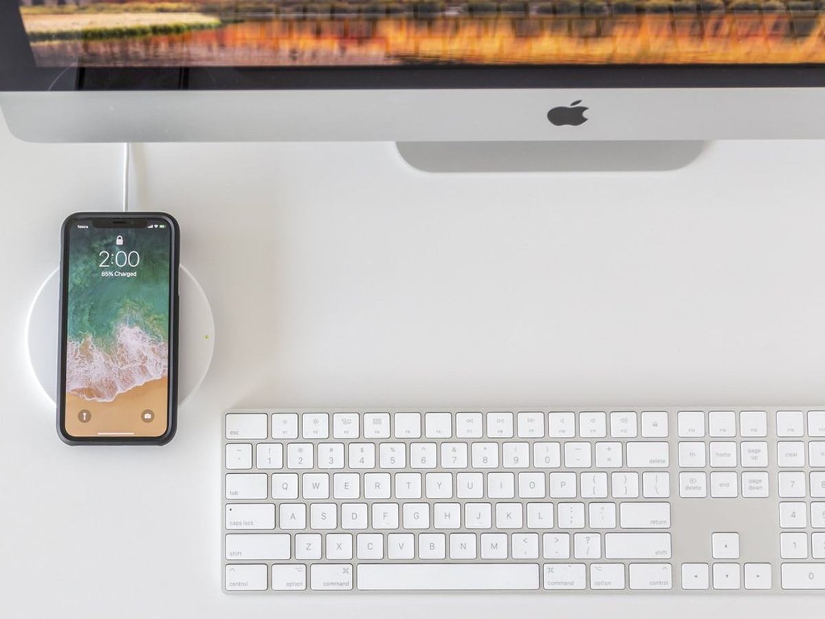 Draufsicht auf ein Telefon, das auf einem weißen kabellosen Ladegerät geladen wird, neben einer Tastatur und einem Monitor.