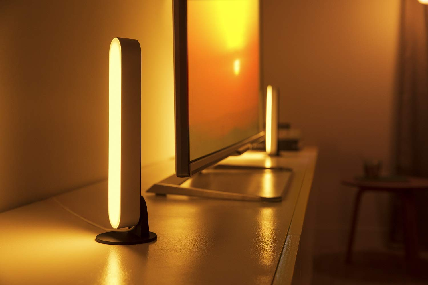 Dos barras de luz Philips Hue Play sobre una superficie blanca junto a un televisor. Iluminación cálida.