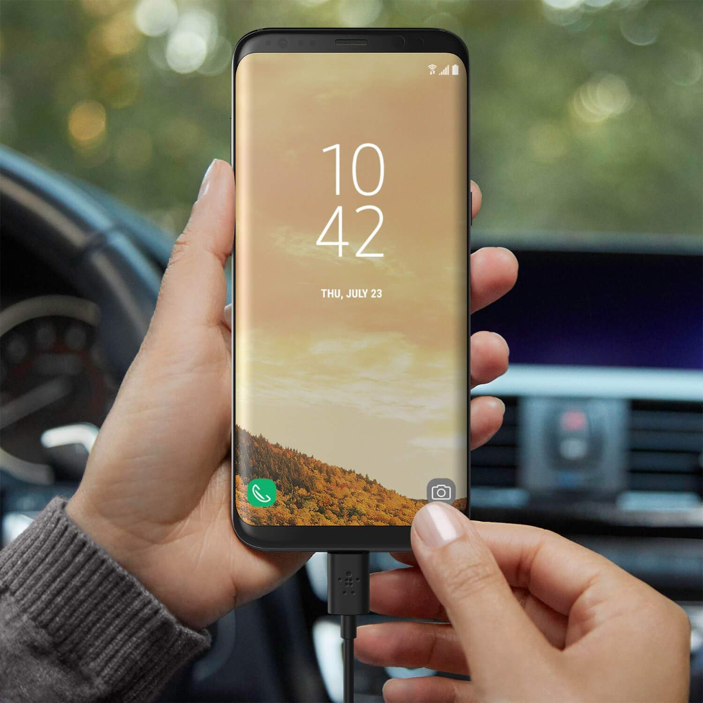 Egy személy egy telefon, ami egy kábelen keresztül töltődik. A képernyő naplementét mutat.