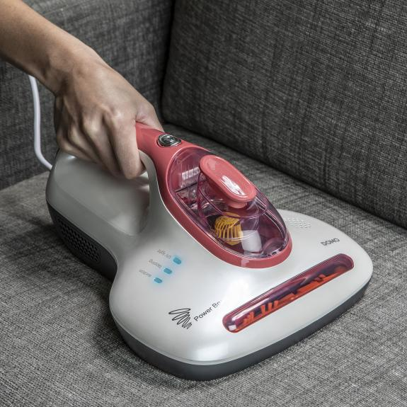 Main tenant un aspirateur à main sur un canapé gris. L'aspirateur est blanc et rouge.