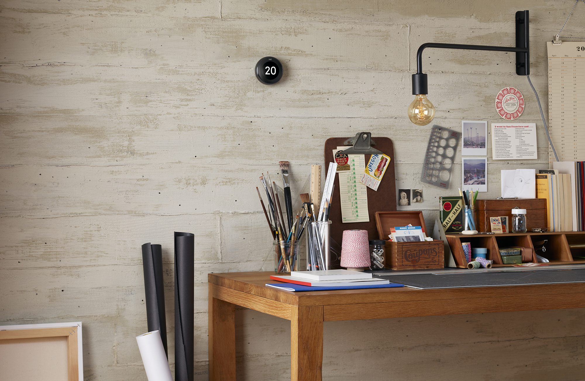 Een bureau met schildersbenodigdheden, een Nest thermostaat en een wandlamp.