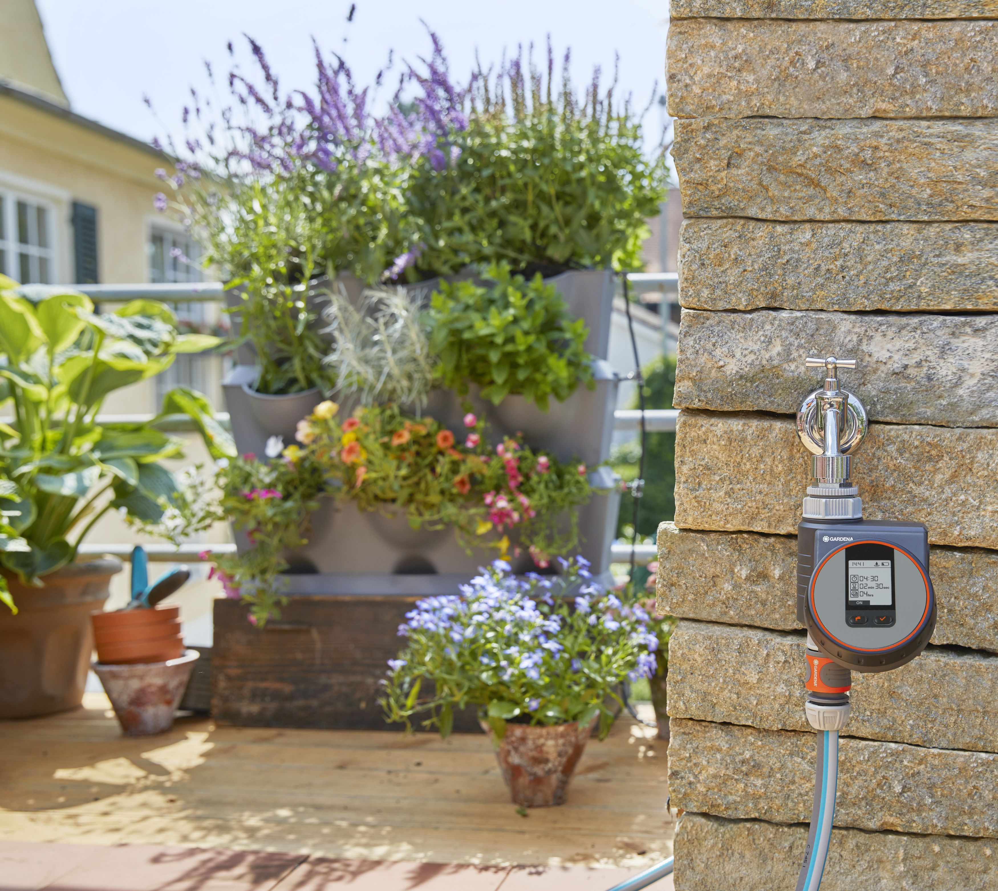 Eine Gartenszene mit Bewässerungssystem, Pflanzen und einer Person auf einer Holzterrasse.