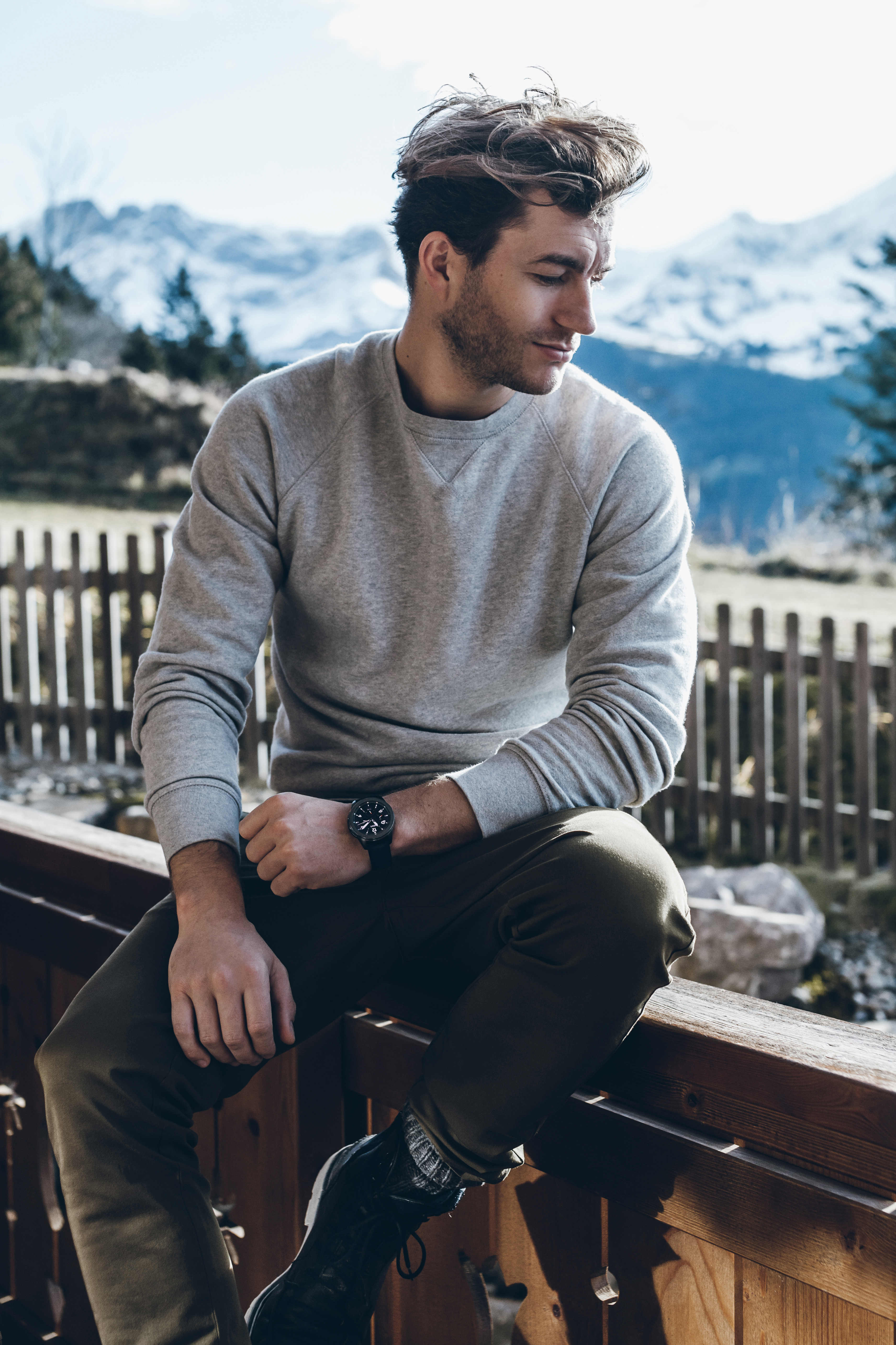 Ein Mann sitzt auf einem Geländer mit Bergkulisse. Er trägt einen grauen Pullover, eine Uhr und eine Hose. Der Himmel ist hell.