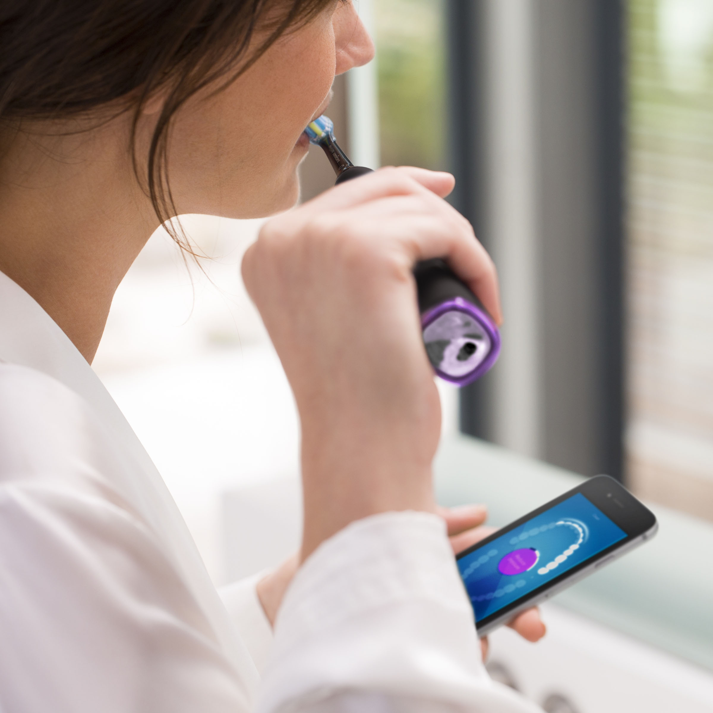 Frau putzt sich mit einer elektrischen Zahnbürste die Zähne und schaut auf ein verbundenes Telefon.