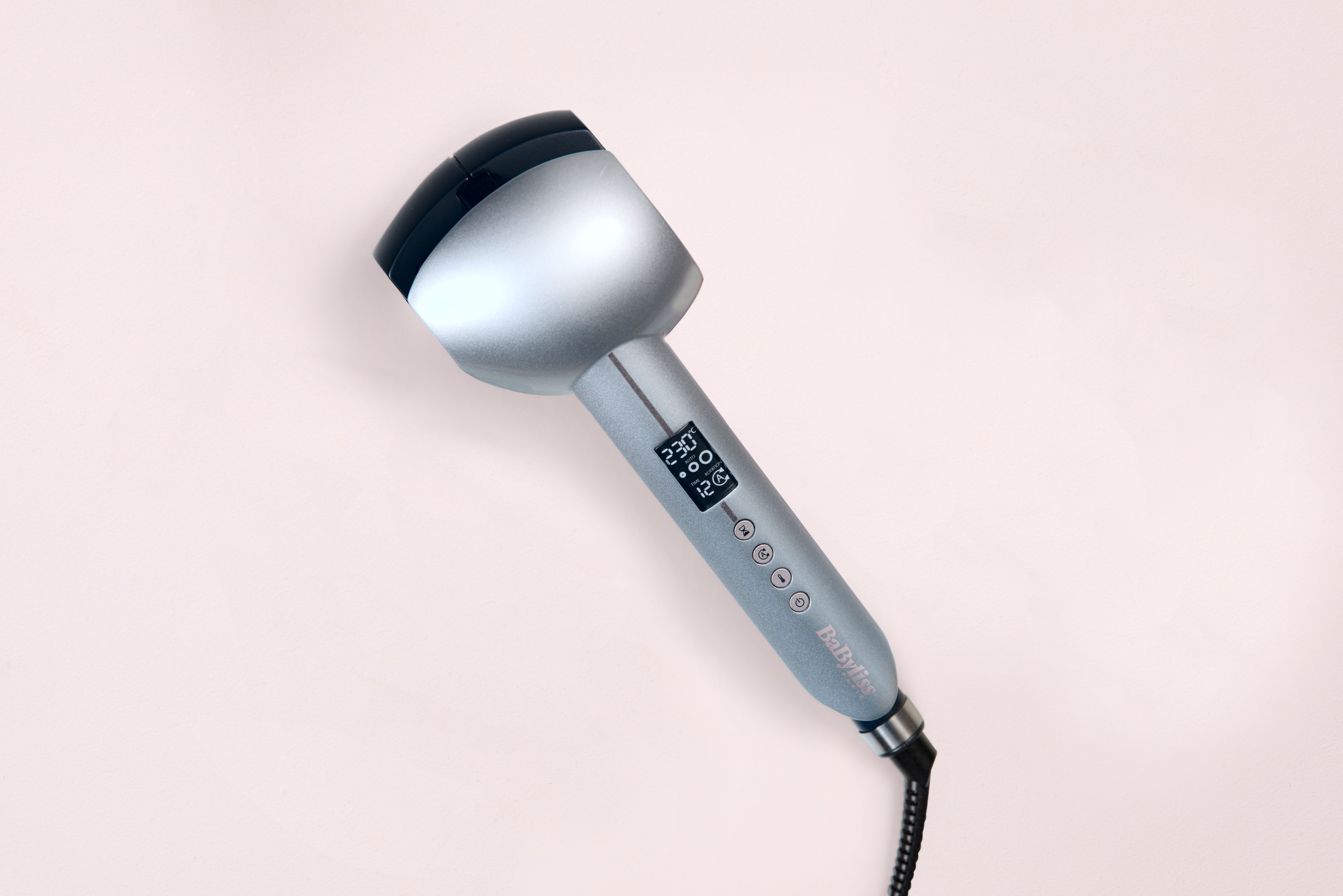 A silver and black curling iron lies on a pink surface. It has a digital display showing settings. The brand name is visible.