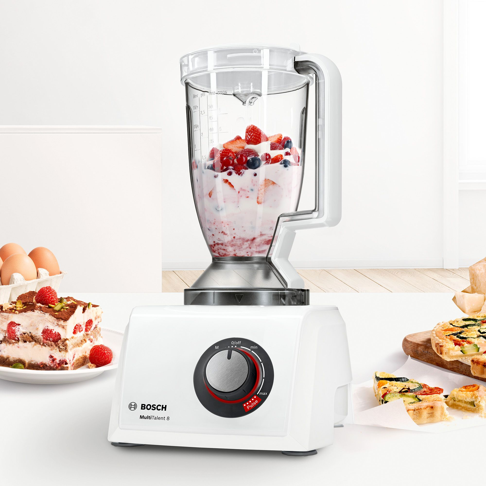 White Bosch blender with fruit smoothie, cake, and quiche on a white surface.