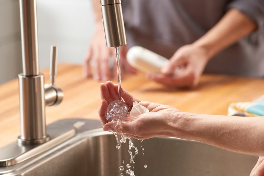 Hände einer Person spülen einen Babyflaschenteil unter fließendem Wasser am Waschbecken ab.