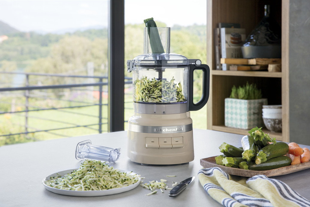Cremefarbener Foodprozessor mit Zucchini. Andere Zucchini und Karotten auf dem Tisch.
