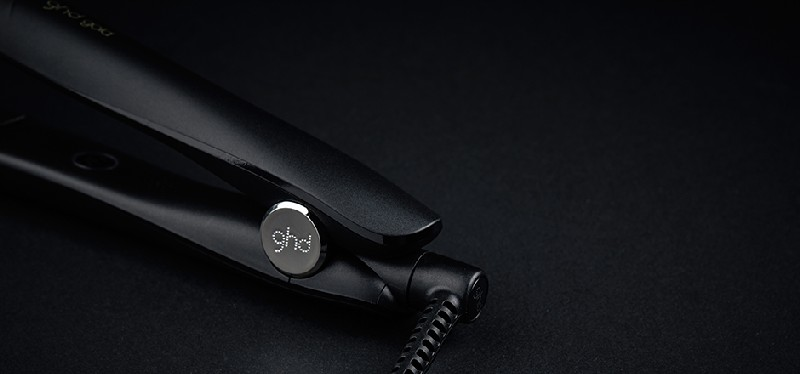 Plancha de pelo negra, vista en ángulo, con el logo GHD sobre una superficie negra.